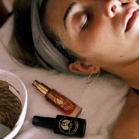 A person lying back on a pale towel, eyes closed, freckled skin bare of makeup, with two Sacred Rituel bottles resting on the towel beside their head: an amber-gold glass bottle, and a black bottle whose gold emblem reads SACRED ROSE MIST.