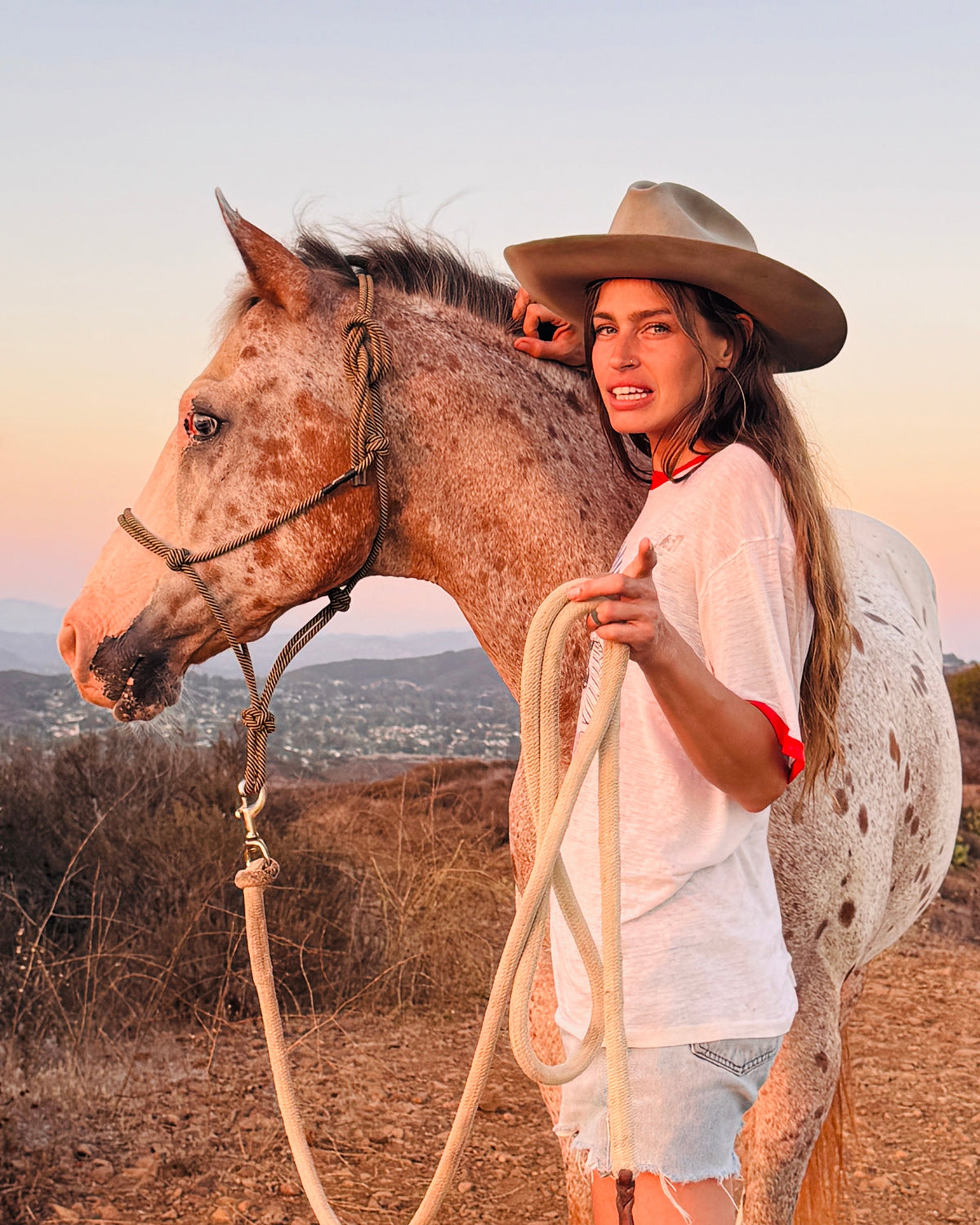 A woman in a cowboy hat standing with her appaloosa horse on a hilltop at dusk, holding its lead rope
