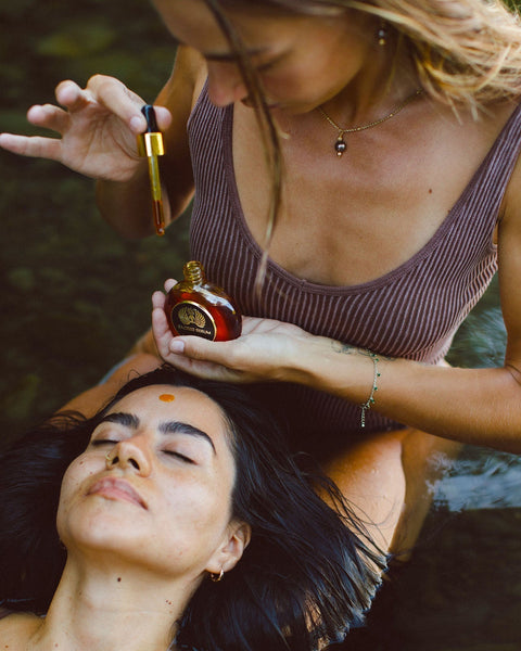 One woman drips golden serum from a dropper onto another woman's forehead as she rests back in natural water, a whole-plant skincare ritual moment