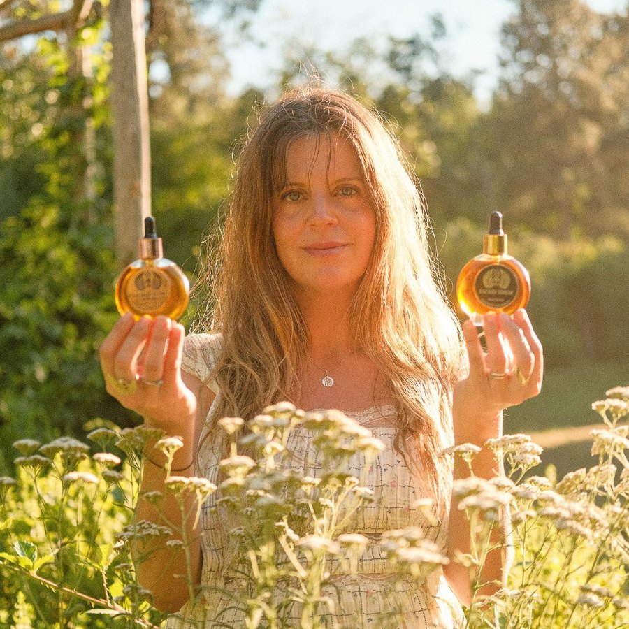 Marysia Miernowska stands in a field of yarrow in golden evening light, holding a bottle of Sacred Rituel oil in each hand